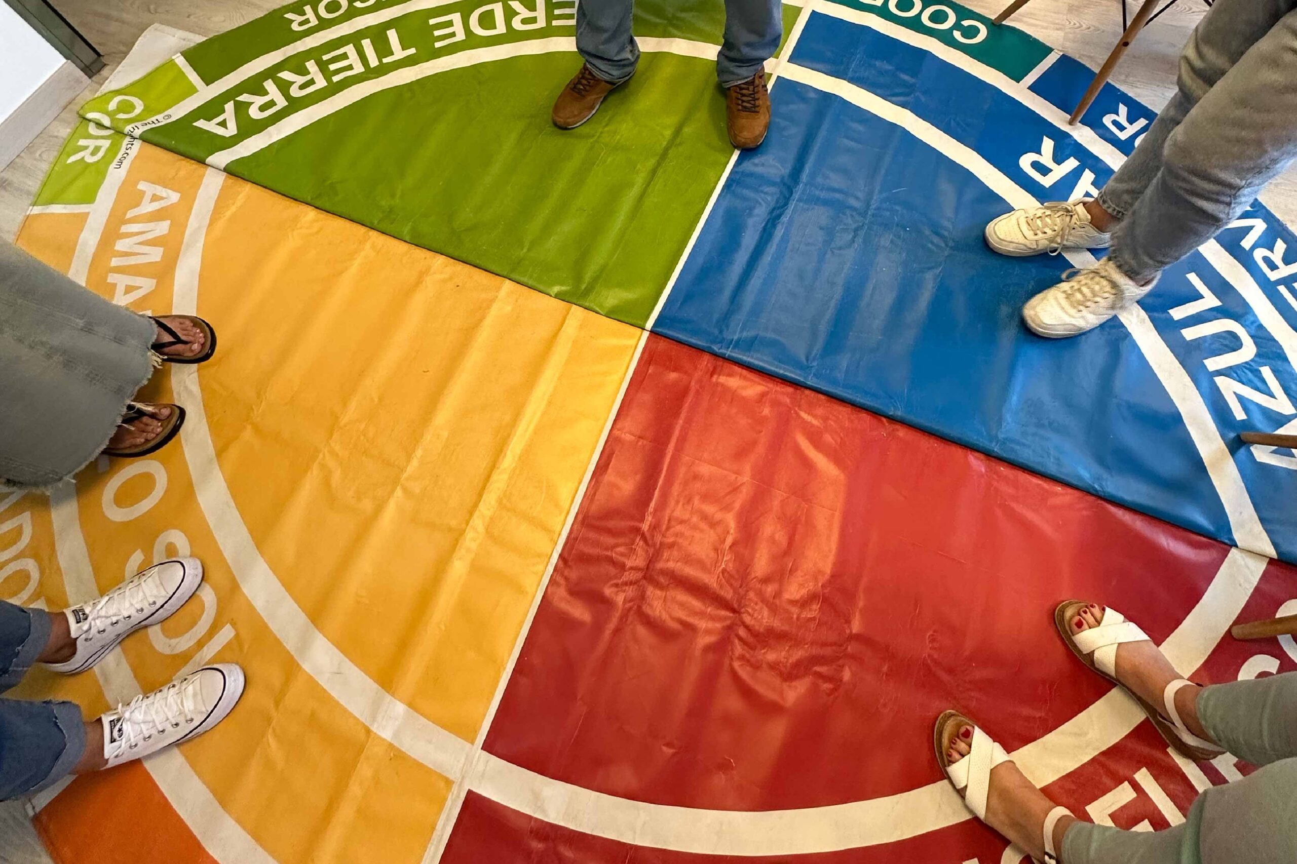 Dinámica de liderazgo basada en los cuatro colores de Insights Discovery Profesional dirigiendo una dinámica grupal en una formación de Insights Discovery, con participantes alrededor del mapa de los cuatro colores en el suelo.